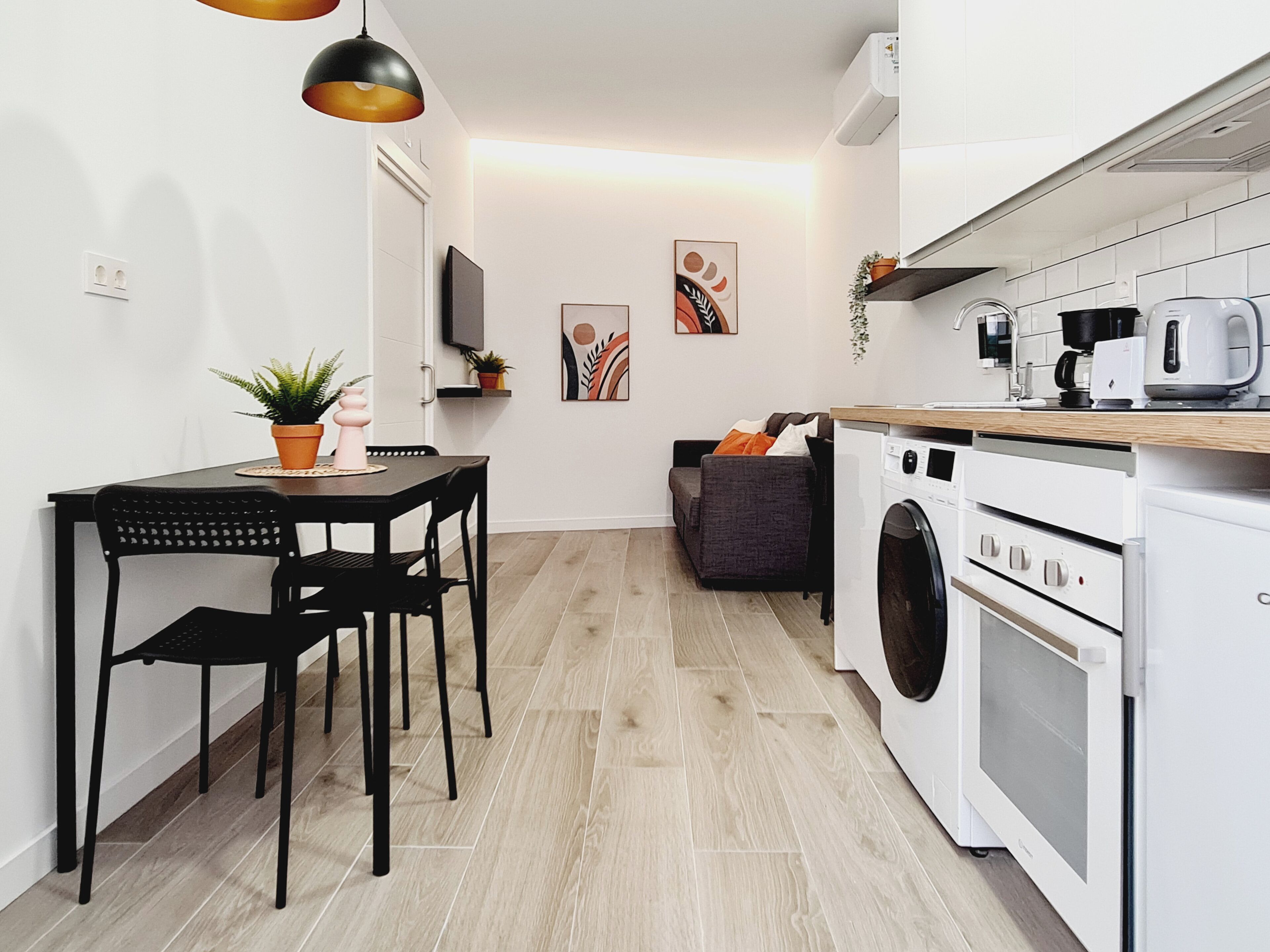 A compact kitchen with a washing machine, a dining table with chairs, a sofa, and a TV mounted on the wall.