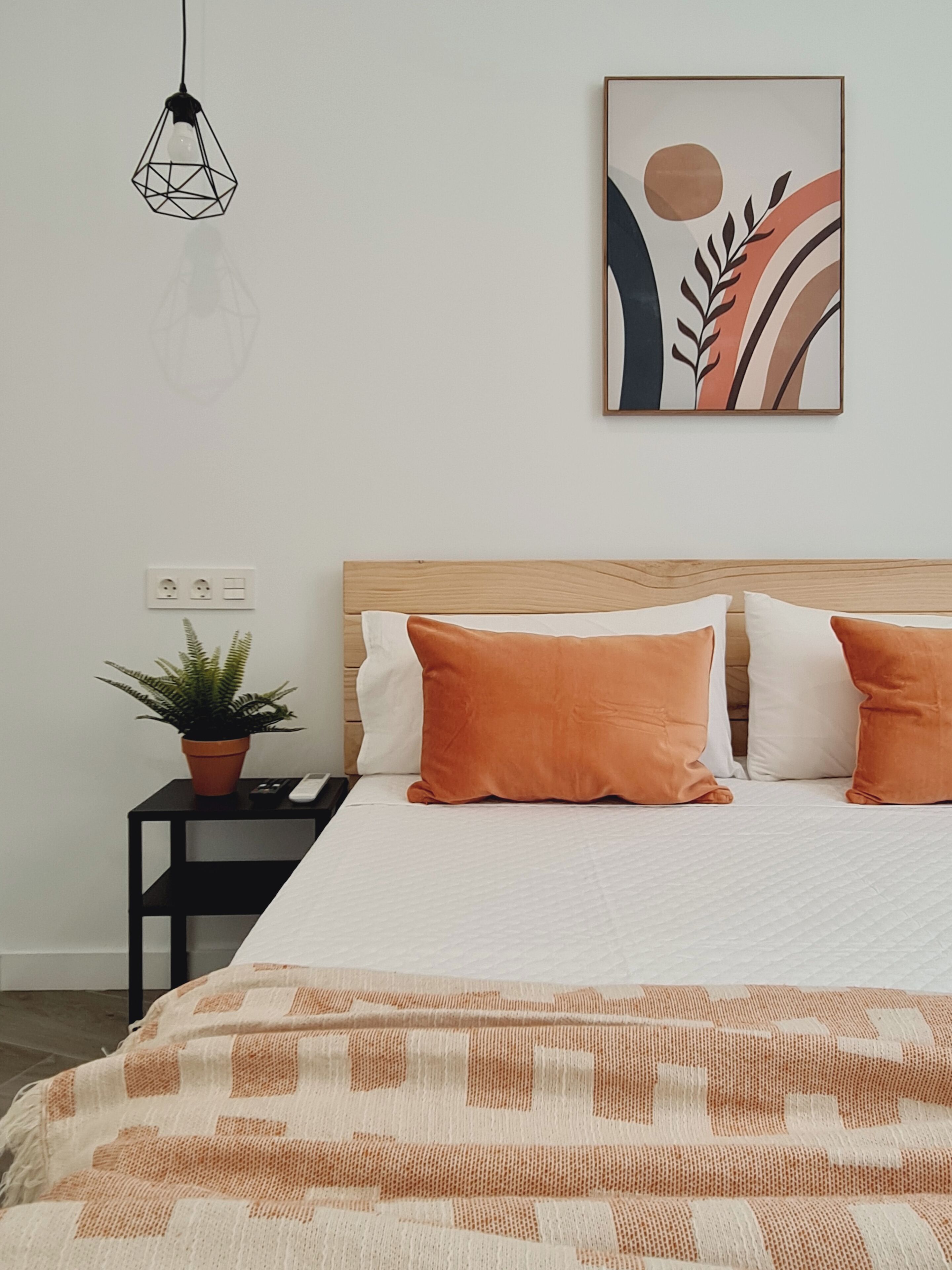A bedroom with a wooden headboard, a white bedspread, two orange pillows, a black nightstand with a potted plant, and a wall-mounted abstract painting.