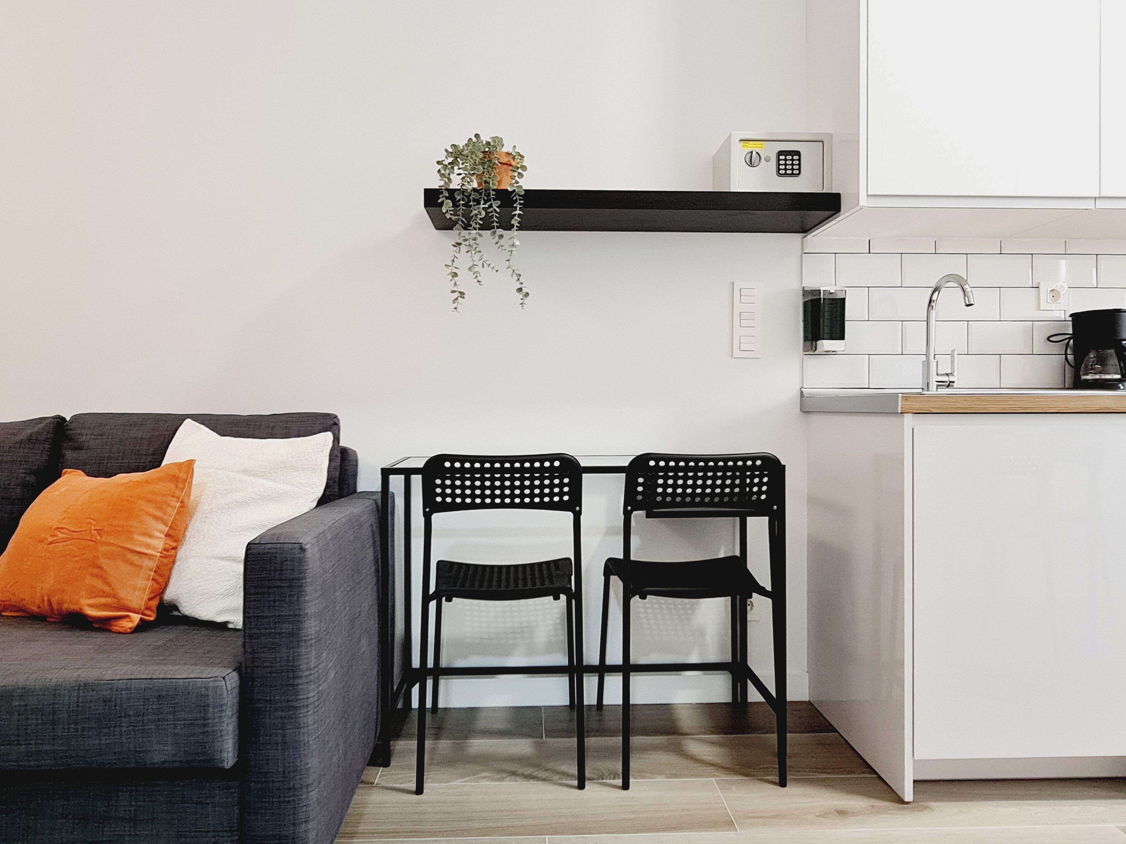 Un espacio habitable compacto con sofá, dos sillones, una mesita, zona de cocina con cafetera y un dispositivo de seguridad en la pared.