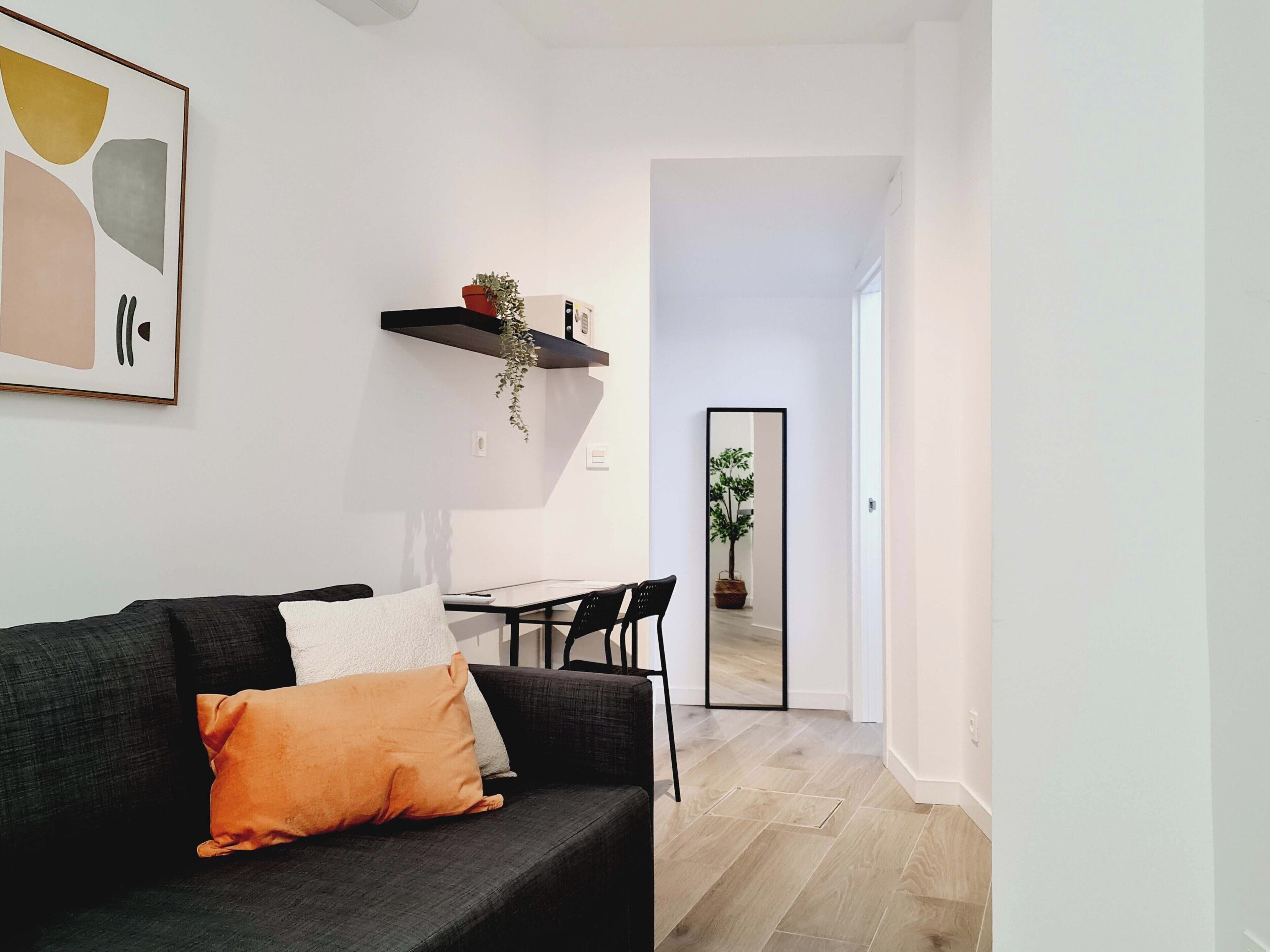 Una sala de estar moderna con un sofá gris oscuro, un cojín blanco y una almohada color mostaza. Una mesa de centro negra con una computadora portátil y una silla negra está en el centro. Una estantería negra con una planta cuelga de la pared. Un espejo grande refleja una planta en maceta y un umbral que conduce a otra habitación.