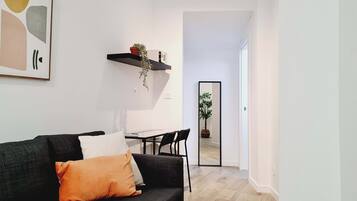 Una sala de estar moderna con un sofá gris oscuro, un cojín blanco y una almohada color mostaza. Una mesa de centro negra con una computadora portátil y una silla negra está en el centro. Una estantería negra con una planta cuelga de la pared. Un espejo grande refleja una planta en maceta y un umbral que conduce a otra habitación.