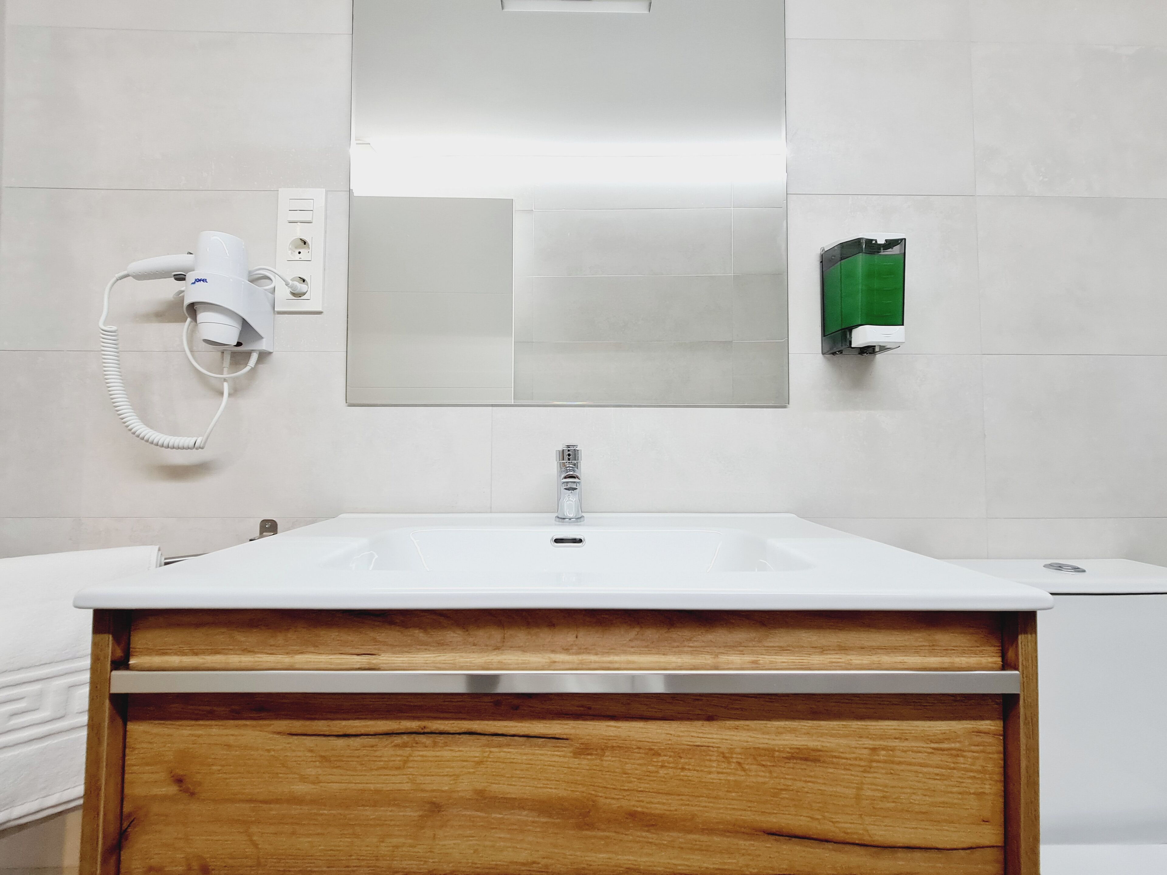 Een badkamer met een witte wasbak, een houten wastafelmeubel, een spiegel, een haardroger en een zeepdispenser.