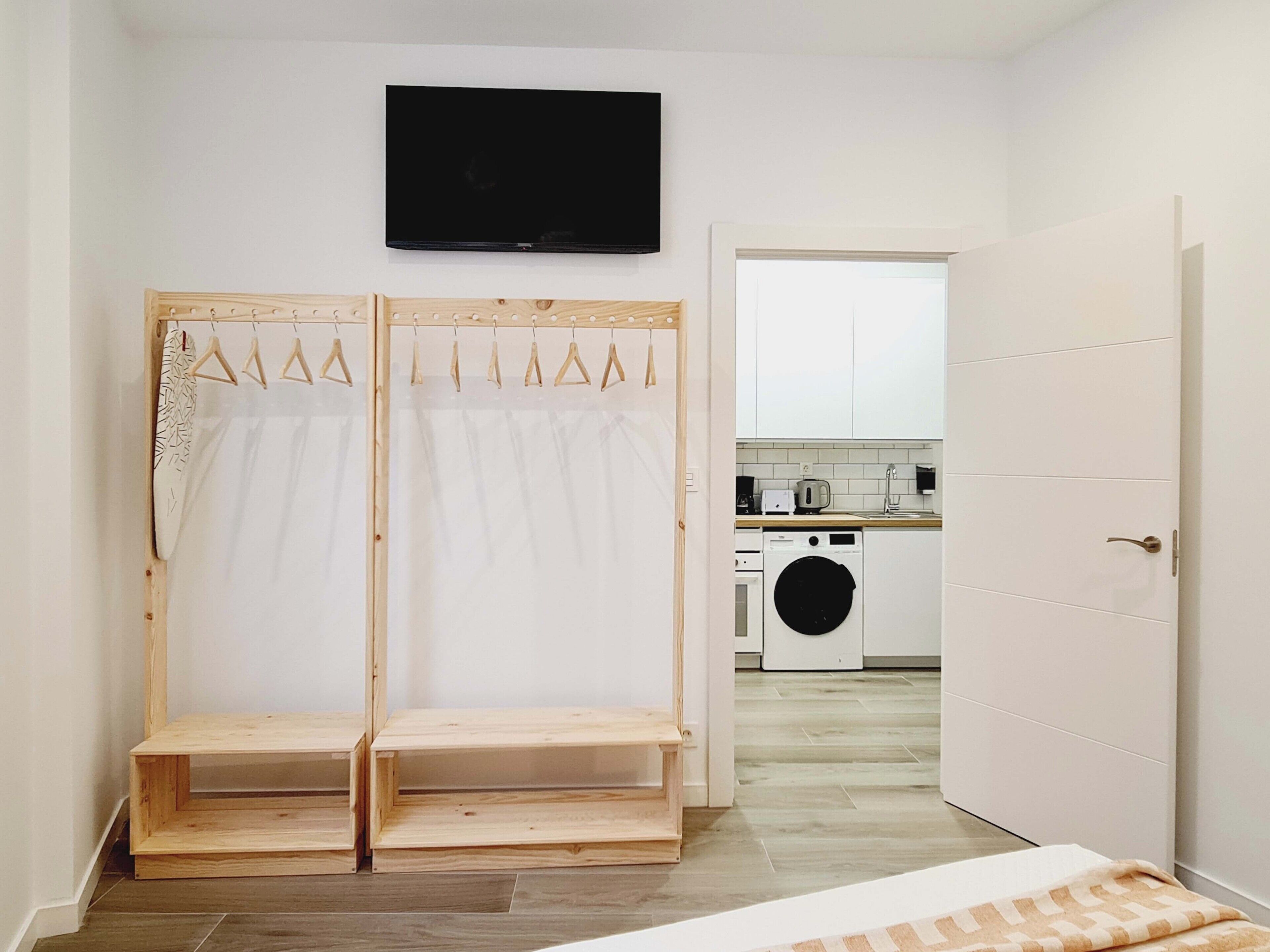 A compact room with a bed, a TV mounted on the wall, a washing machine, and a small kitchenette.