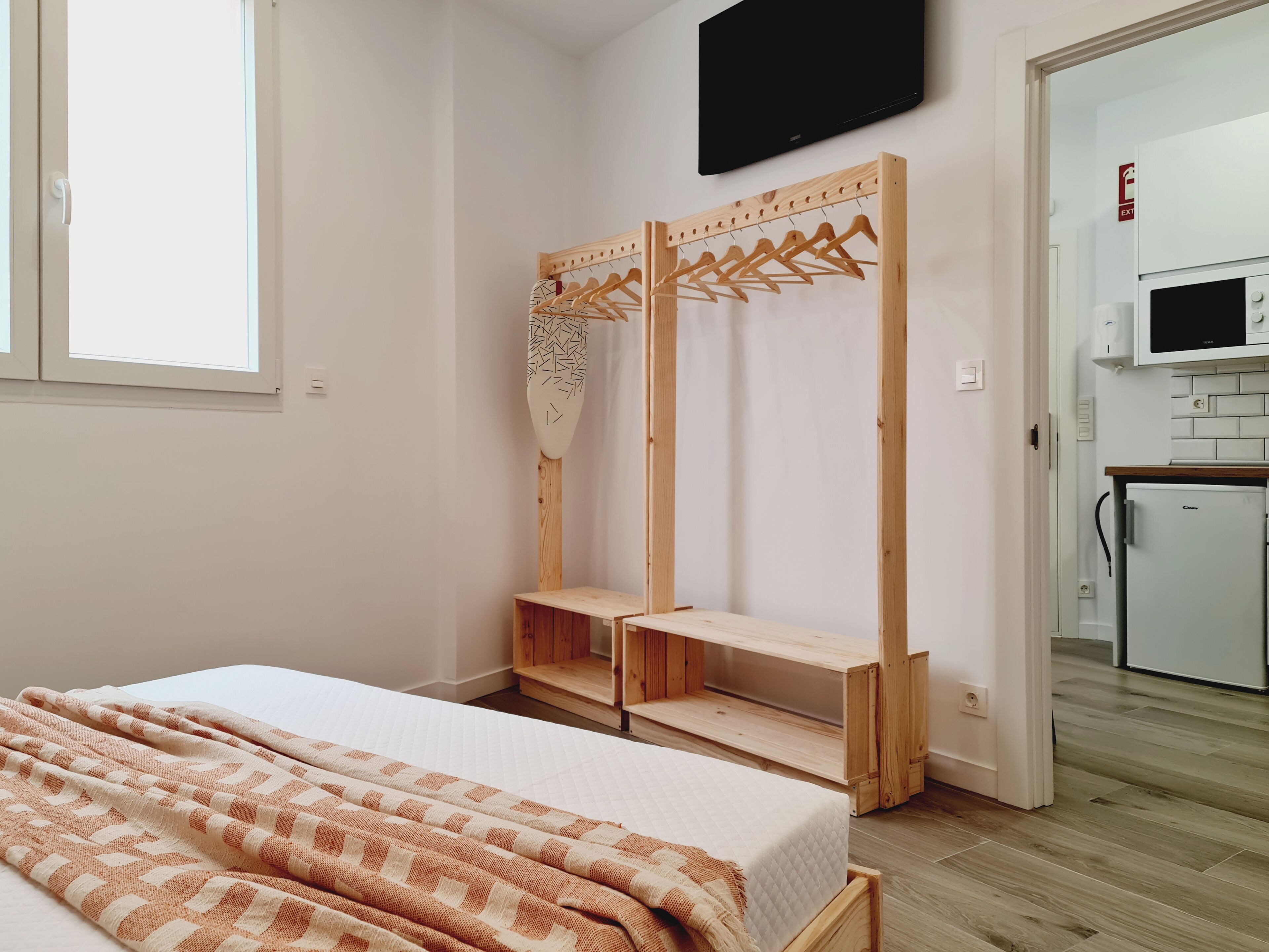 A bedroom with a bed, a wooden wardrobe, a television, and a kitchenette.