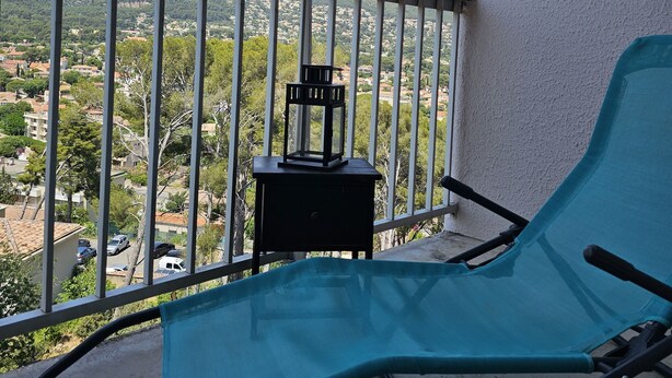 A balcony with a blue chair, a small table with a lantern, and a view of a mountainous landscape. - Appartement Cosy 4 Personnes (Toulon)