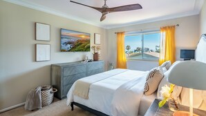 A bedroom with a bed, a nightstand, a ceiling fan, a window with curtains, and artwork on the wall.