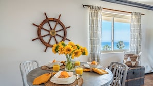 A dining area with a round table, chairs, a ship's wheel decoration, a vase of sunflowers, and a view of the ocean through a window with patterned curtains.