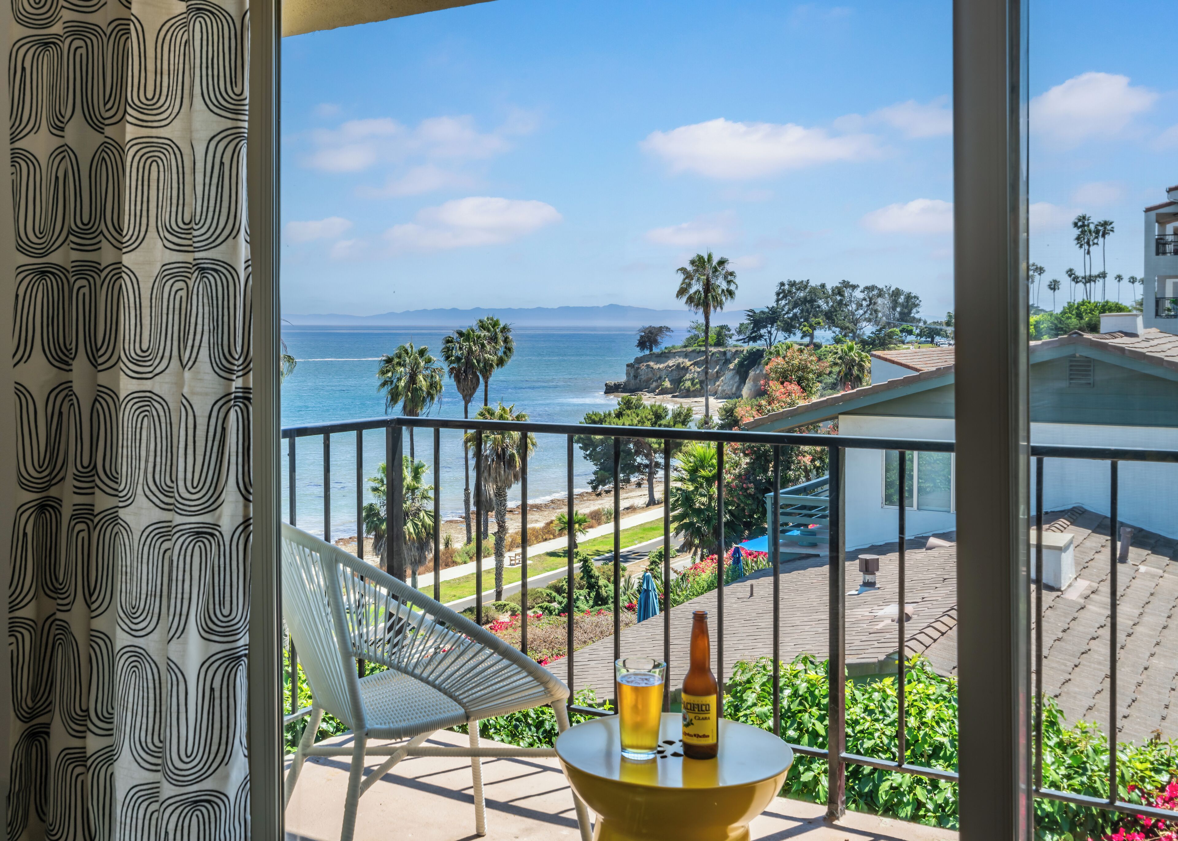 Uma varanda com vista para o oceano, uma cadeira, uma mesa com uma garrafa e um copo, e uma cortina estampada.