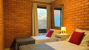 Una habitación con paredes de ladrillo, dos camas con almohadas rojas y blancas, una mesita de noche con lámpara y una ventana con cortinas.