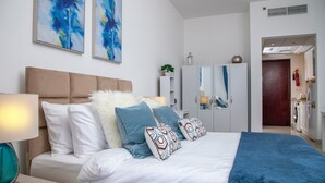 Un dormitori modern amb un llit gran, roba de llit blanca i almoinades decoratives. Hi ha una tauleta de nit amb una làmpada i un armari amb un mirall. Es veu un armari i una porta que condueix a un bany al fons.