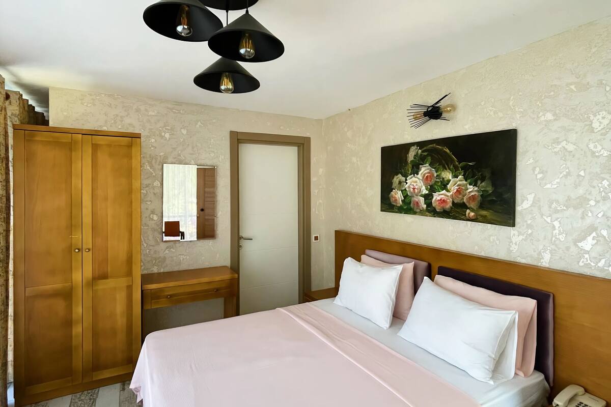 A bedroom with a bed, bedside table, nightstand, and a telephone. There is a wardrobe, a mirror, and a painting on the wall.
