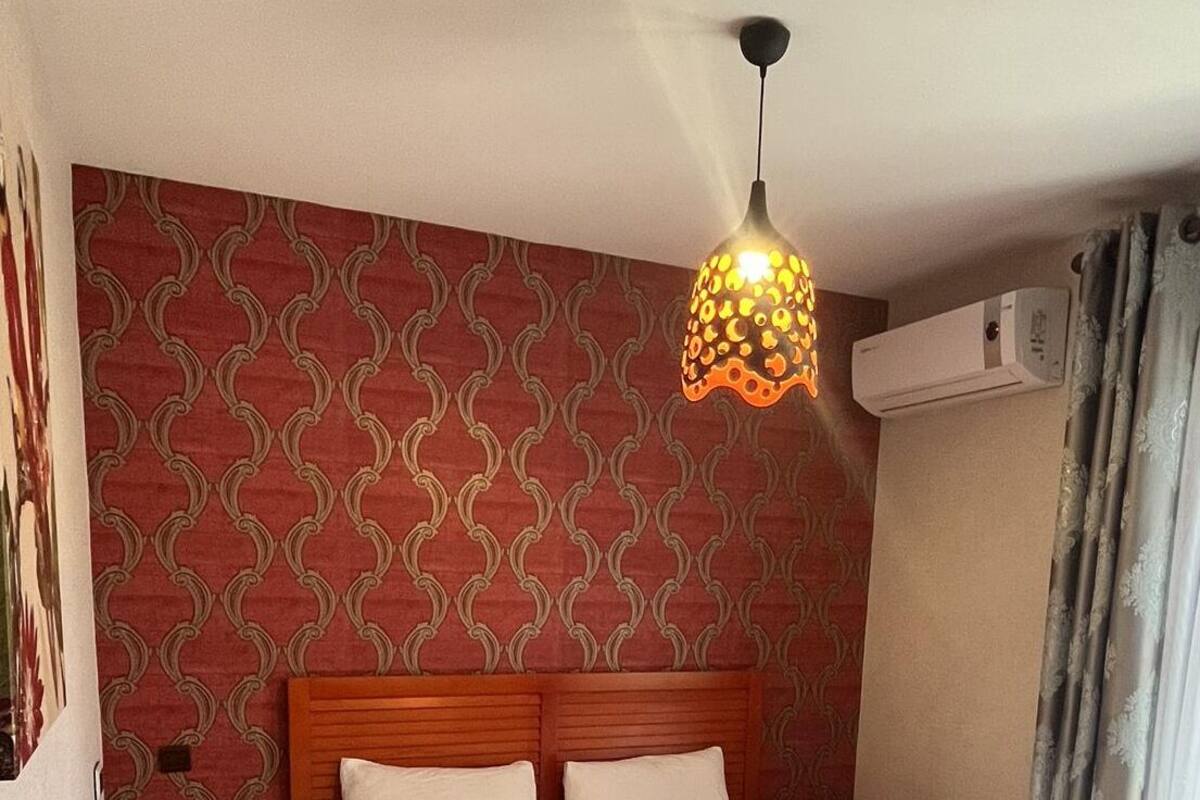 A hotel room with a patterned wallpaper, a wooden headboard, a bed with white linens, a bedside table, and a hanging light fixture.