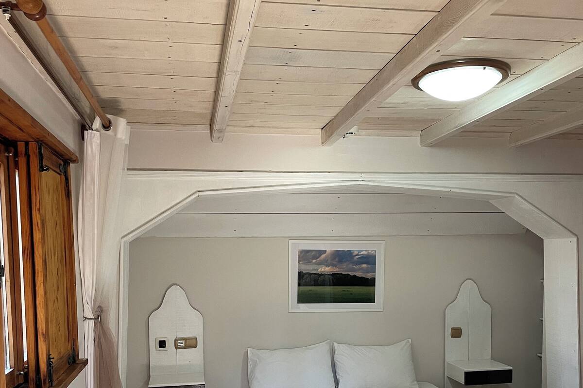 A bedroom with a single bed, wooden ceiling beams, a window with curtains, a wall-mounted light fixture, and a framed picture.