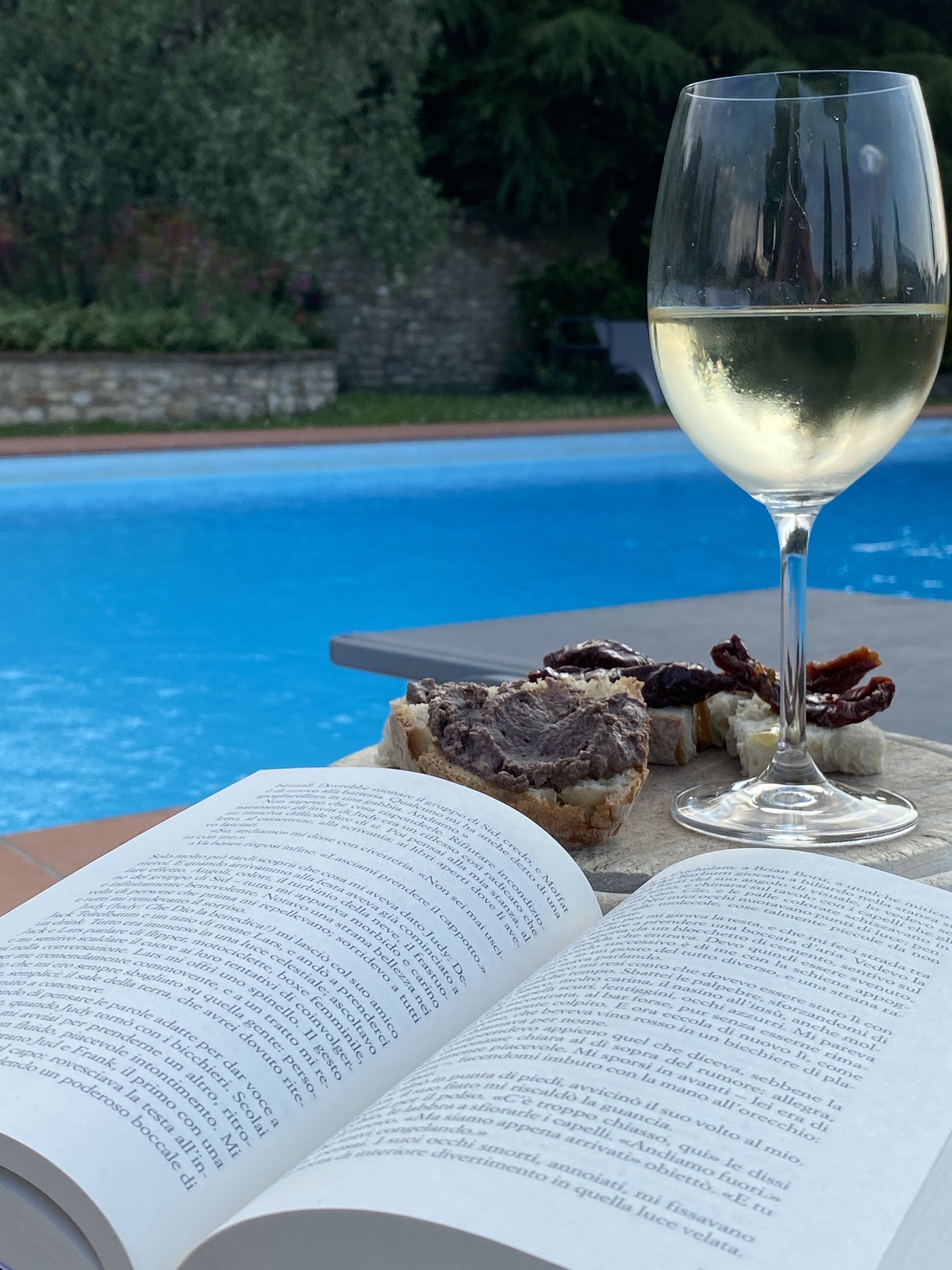 An open book on a table with a glass of white wine and some food items.