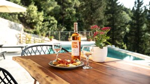 A wooden table with a bottle of wine, two glasses, a plate of food, and a potted plant by a poolside. - Kipseli Villa, Overlooking Vassiliki bay (Lefkada)