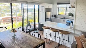 Eine moderne Küche mit einer großen Kochinsel und Barhocker, ein Essbereich mit Holztisch und Stühlen sowie große Fenster mit Blick ins Freie.