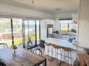 A modern kitchen with a large island and bar stools, a dining area with a wooden table and chairs, and large windows offering a view of the outdoors. - The Golden Retreat @ Hyams Beach - Pet Friendly, Linen & Wifi provided (Hyams Beach)