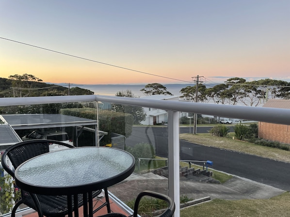 A balcony with a glass table and chairs, overlooking a residential street and a body of water during sunset.