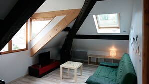 Habitación estilo loft con techo inclinado, un sofá verde, una mesita, un televisor y una lámpara.