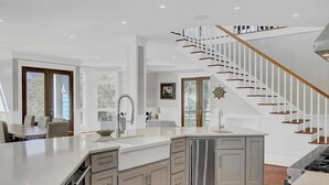 A modern kitchen with a central island, a built-in dishwasher, and a staircase with wooden railings. - Second Star | Oceanview Coastal Showpiece with Private Pool (Isle of Palms)