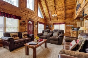 A cozy cabin interior with wooden walls, a fireplace, a dining area, and a living room with leather sofas and a coffee table.