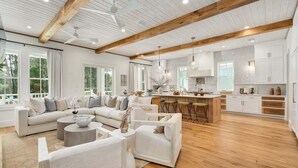 A spacious living area with a large L-shaped sofa, a coffee table, and a kitchen with white cabinets and wooden beams.