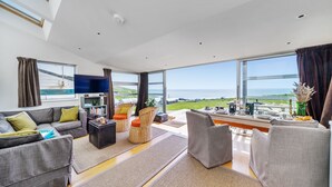 A spacious living room with a large window offering a view of the sea, a flat-screen TV mounted on the wall, and a comfortable seating area with sofas and a coffee table. - The Billy Shed  - Eype, Bridport (Bridport)
