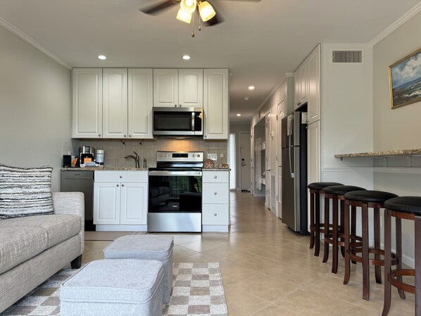 A modern kitchen with white cabinets, stainless steel appliances, and a built-in oven. There is a gray sofa and ottomans in the living area, and wooden bar stools along the kitchen counter.