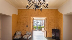 A spacious room with a large chandelier, two patterned armchairs, a wooden cabinet, and a view through an open doorway to a balcony with a railing and greenery. - Seclude Shimla Taraview (Shimla)