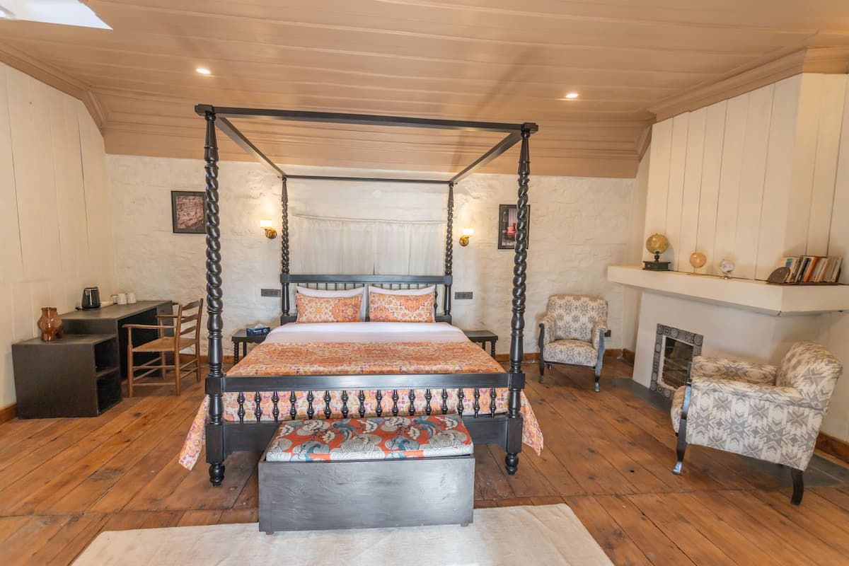Een kamer met houten vloeren en witte wanden, met een hemelbed, een bankje, een open haard en een bureau met stoelen.