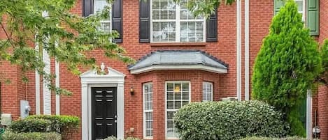A two-story brick house with a front porch, a black door, and a small front yard with bushes and a tree.