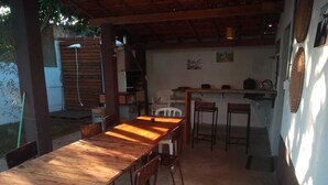 Un espace extérieur abrité, aménagé pour les repas, avec une longue table et des chaises en bois, un petit coin cuisine équipé d’une cuisinière et de tabourets de bar, et un sol en béton.