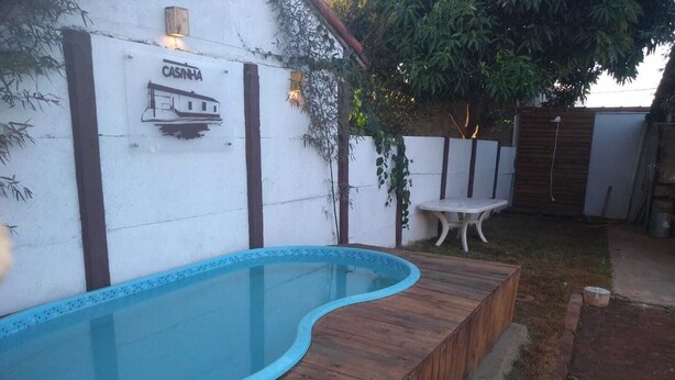 Une piscine avec une terrasse en bois, un mur blanc orné d’une enseigne et une petite table.