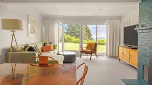 A modern living room with a sofa, ottoman, dining table, and chairs. There is a television on a cabinet and a large window with curtains.