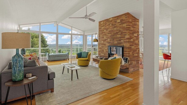 A spacious living room with a fireplace, large windows, and a high ceiling fan.