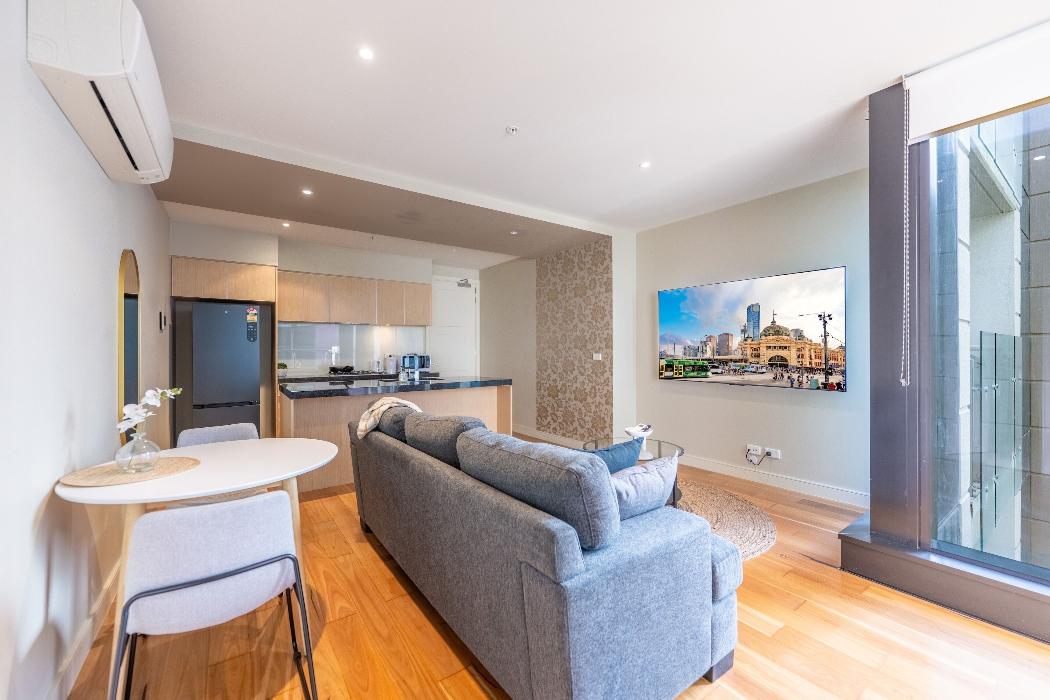 A modern living room with a grey sofa, a dining table with chairs, and a kitchen in the background.