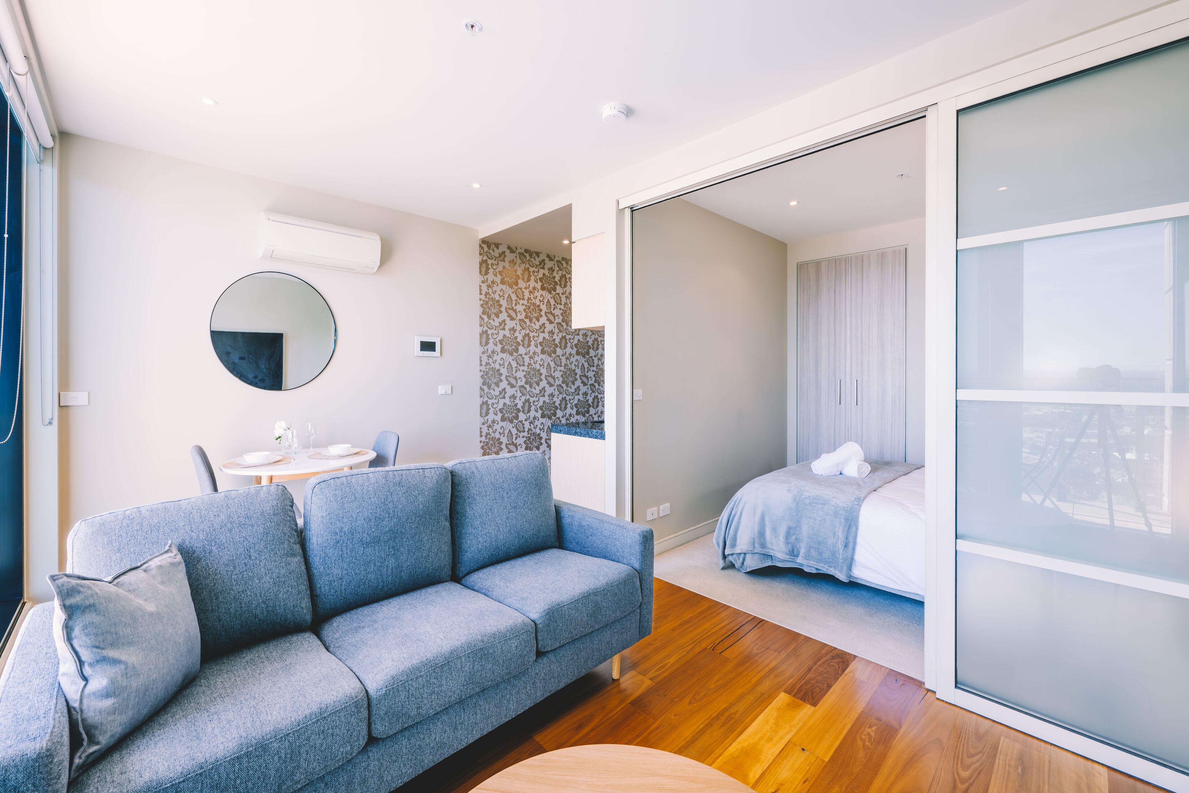 Una habitación de hotel moderna con un sofá gris, un espejo redondo, una mesita redonda y una cama con colcha azul.