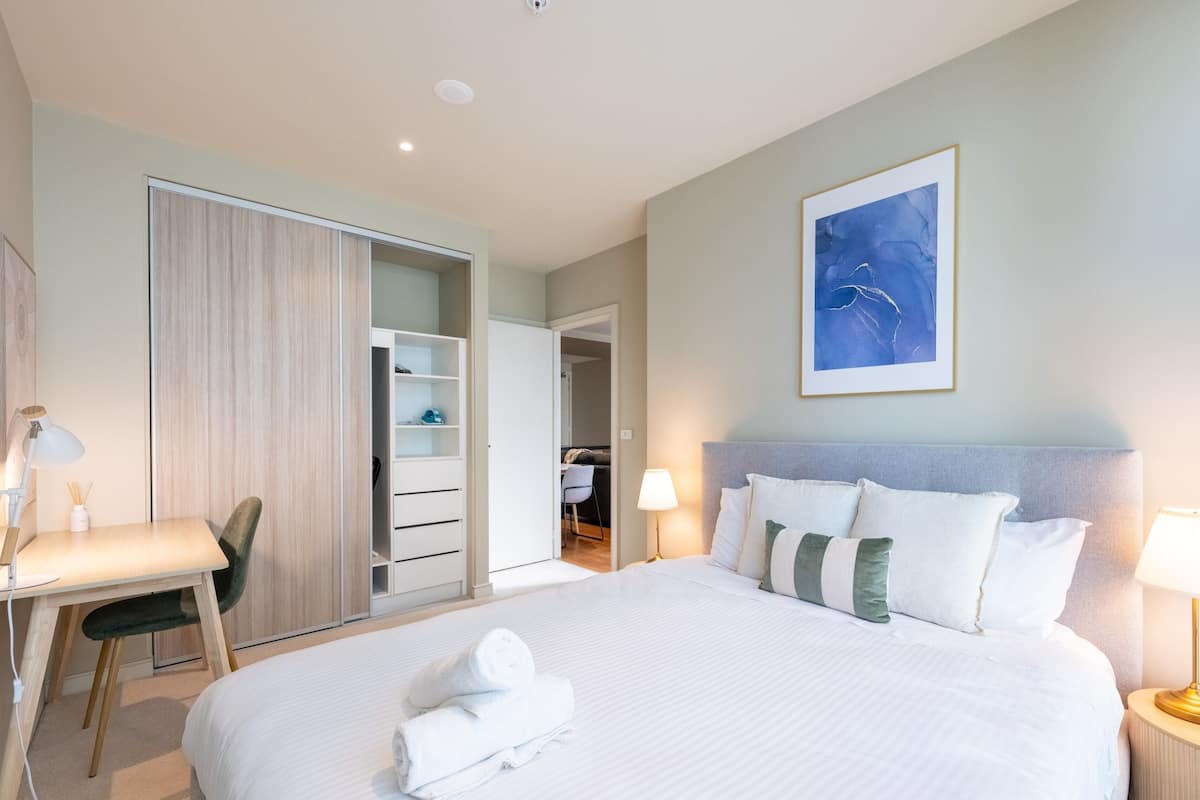 Habitación de hotel con cama, mesita de noche, lámpara, silla, armario y un cuadro en la pared.