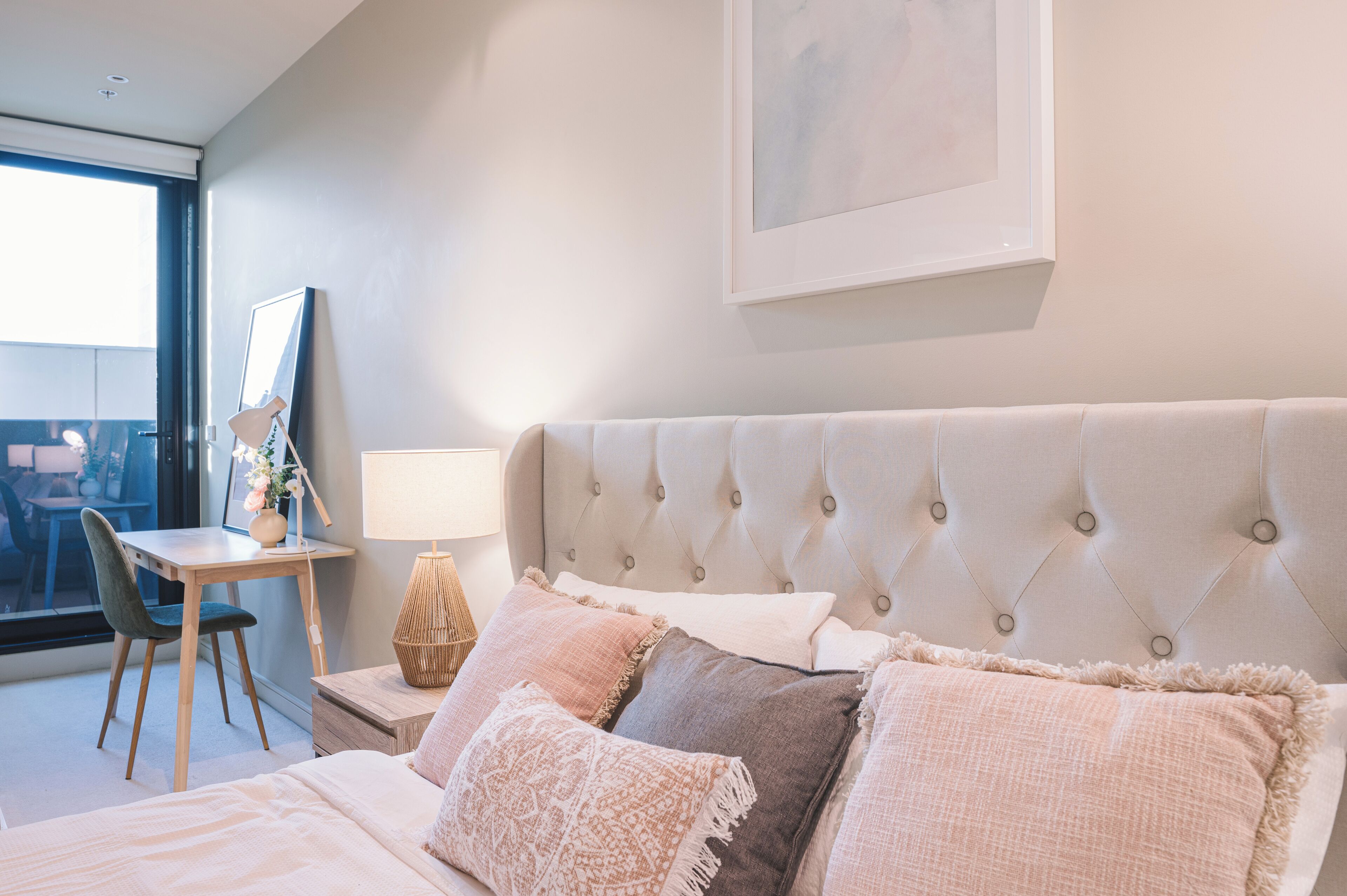 A bedroom with a tufted headboard, a desk with a chair, and a bedside table with a lamp.