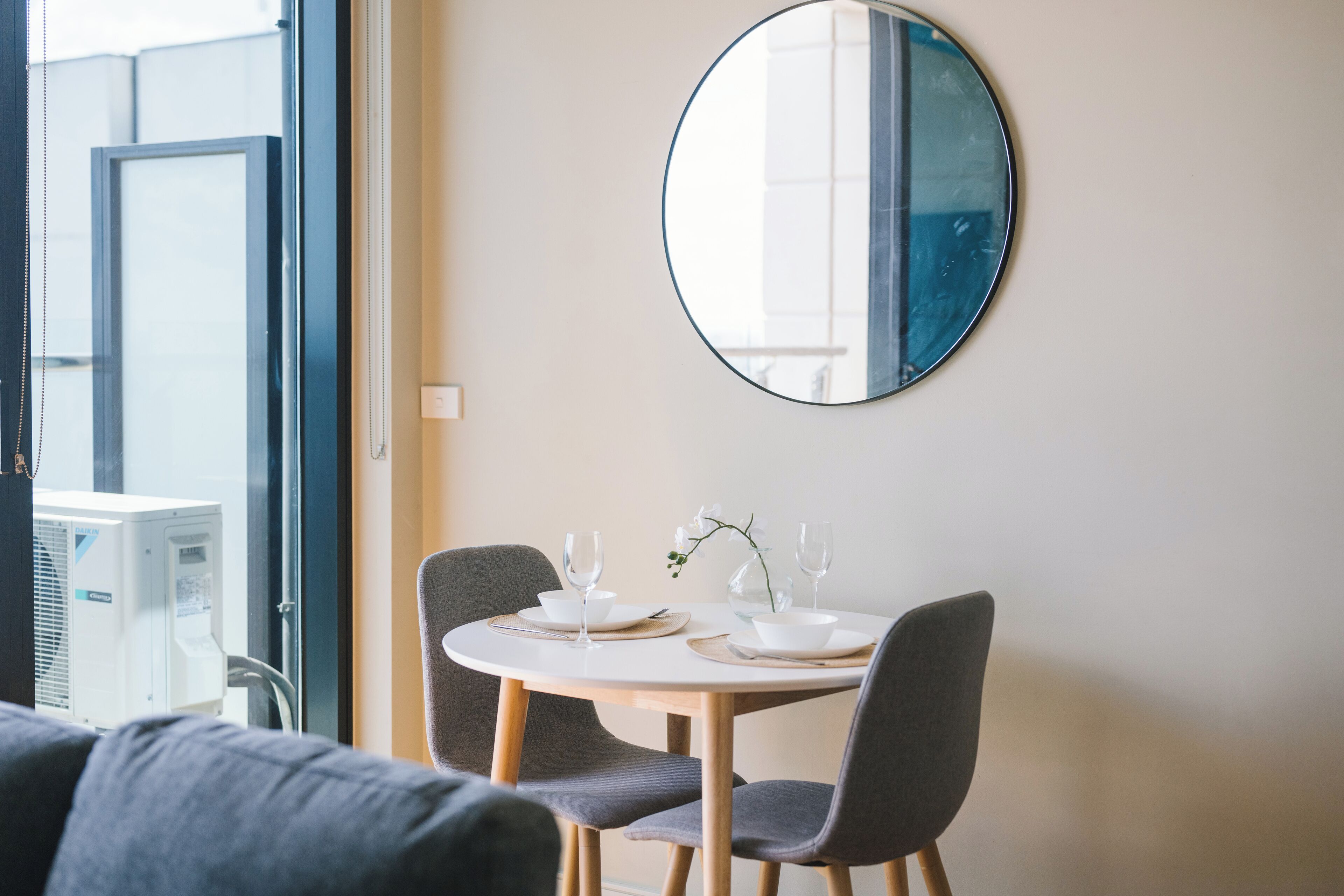 A dining area with a round table set for two, two chairs, a round mirror on the wall, and a window with blinds.