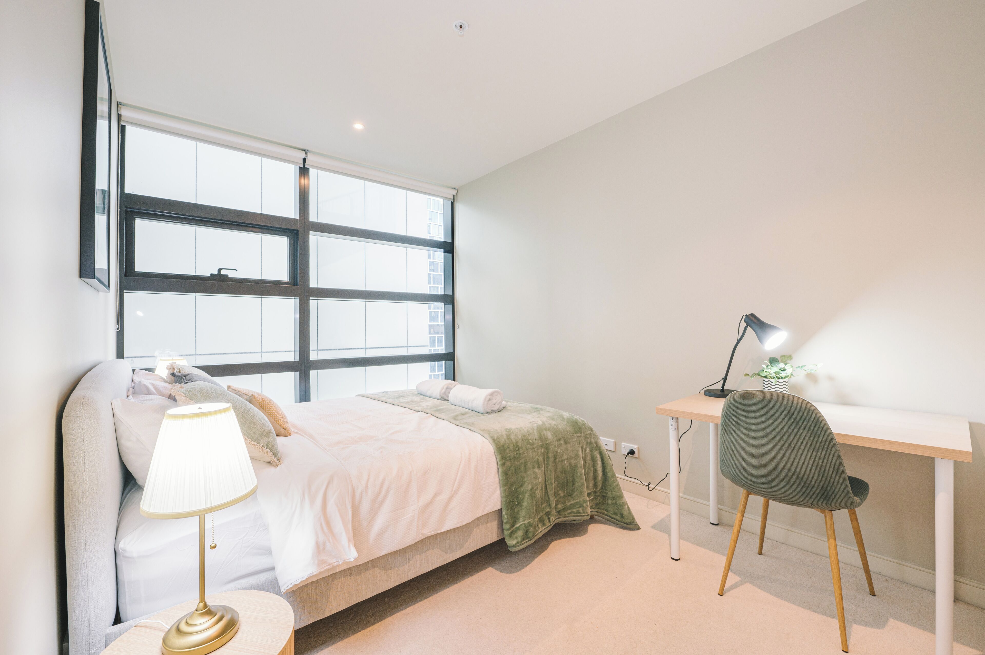 Une chambre à coucher comprenant un lit, une lampe de chevet, un bureau, une chaise et de grandes fenêtres.