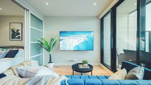Ein modernes Wohnzimmer mit einem großen Fernseher, einem blauen Sofa, einem kleinen Tisch mit Pflanze und einer Glastür, die zu einem Außenbereich führt.