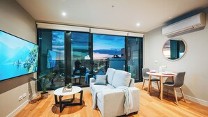 Ein modernes Wohnzimmer mit einem großen Fernseher, einer Couch, einem Esstisch mit Stühlen und Blick auf eine Stadtlandschaft durch große Fenster.