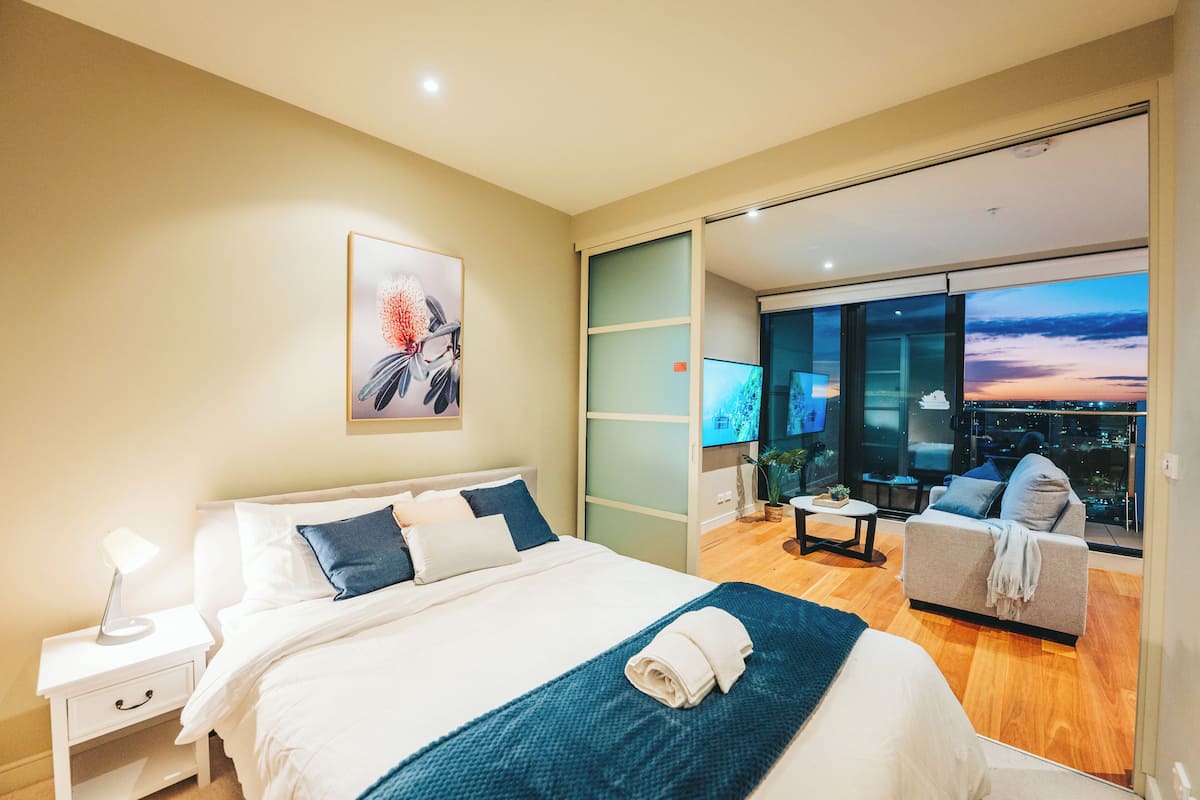 Un dormitorio moderno con una cama grande, una mesita de noche, un televisor y un sofá con vista a la ciudad.