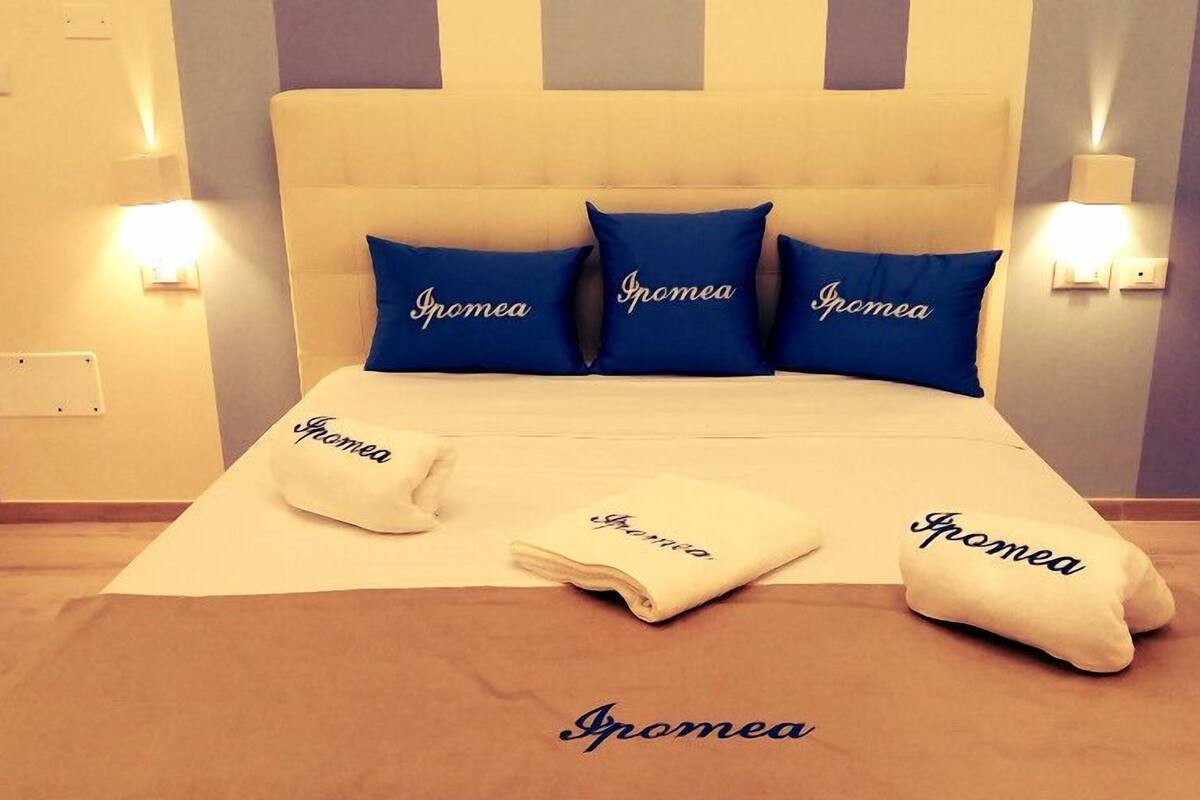 A neatly made bed with blue and white pillows labeled "Ipomea," a matching pillow on the bed, and a matching pillow on the floor.