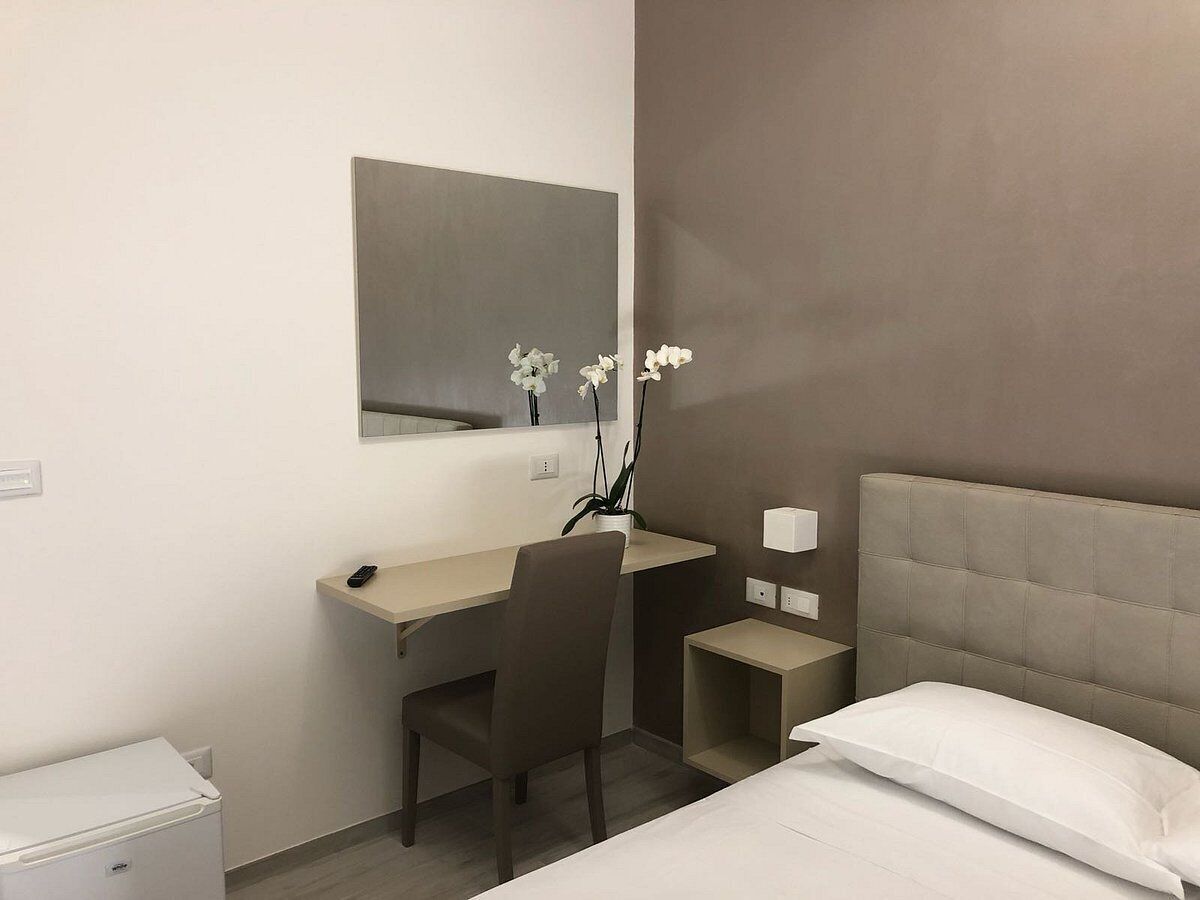 A hotel room with a bed, a desk, a chair, a small bedside table, a mirror, and a potted plant.