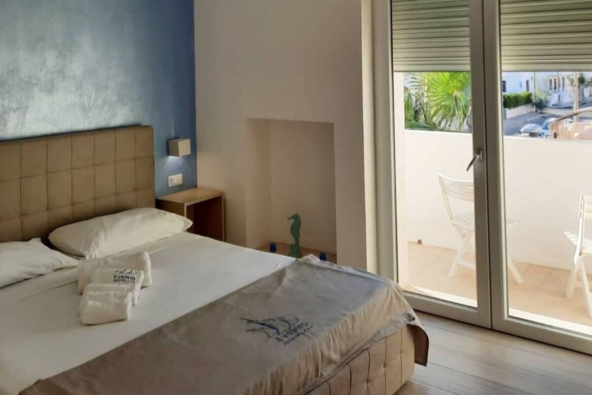 A modern hotel room with a large bed, a ceiling light, a sliding glass door to the balcony, and a view of the outside.