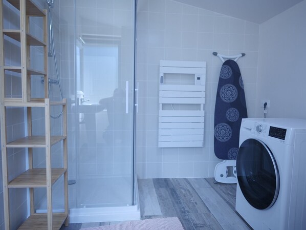 A modern bathroom with a glass shower enclosure, a built-in wooden shelf unit, a white towel rack, and a front-loading washing machine.