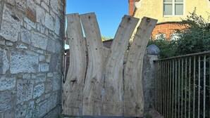 Un portón de madera rústico con un muro de piedra a un lado y una cerca de metal al otro, que da a una casa con techo a dos aguas y chimeneas.