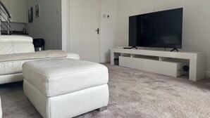 Una sala de estar moderna con un sofá blanco, un reposapiés a juego, un televisor plano grande sobre un mueble blanco y piso alfombrado.